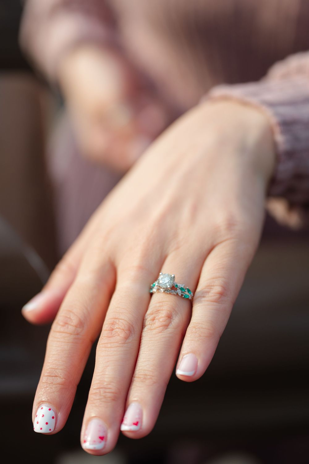 Close-up of a hand displaying a diamond engagement ring with blue stones and heart-patterned French manicure nails.
