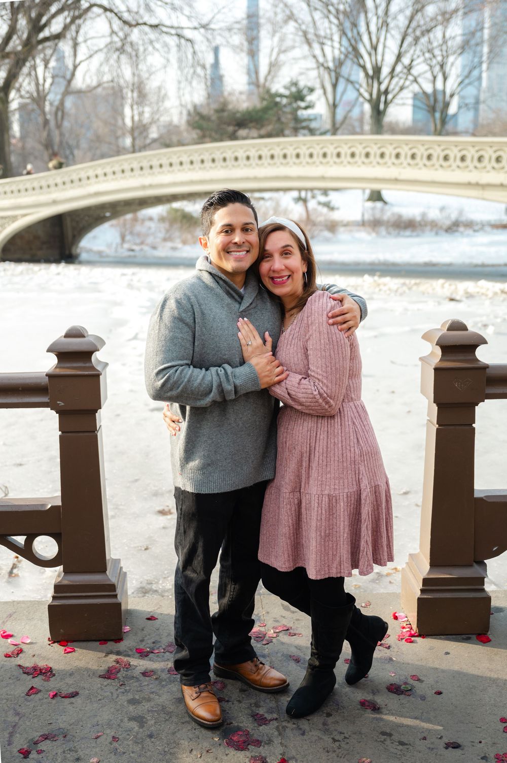 Happy couple embracing at Bow Bridge in Central Park during winter, surrounded by rose petals on the ground.