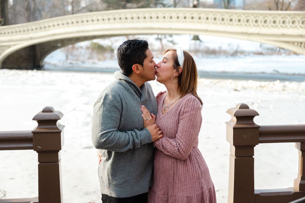Happy engaged couple kissing near iconic Bow Bridge in Central Park during a snowy winter day.