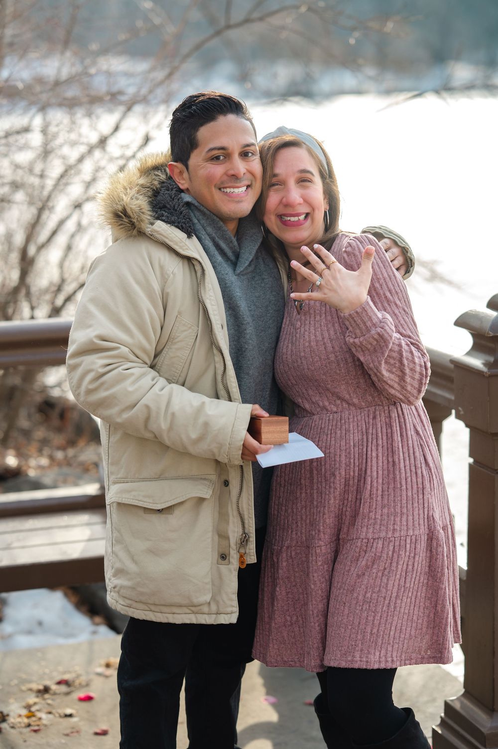 Happy couple on a snowy bridge showing engagement ring, man holding ring box, both smiling outdoors in winter coats.