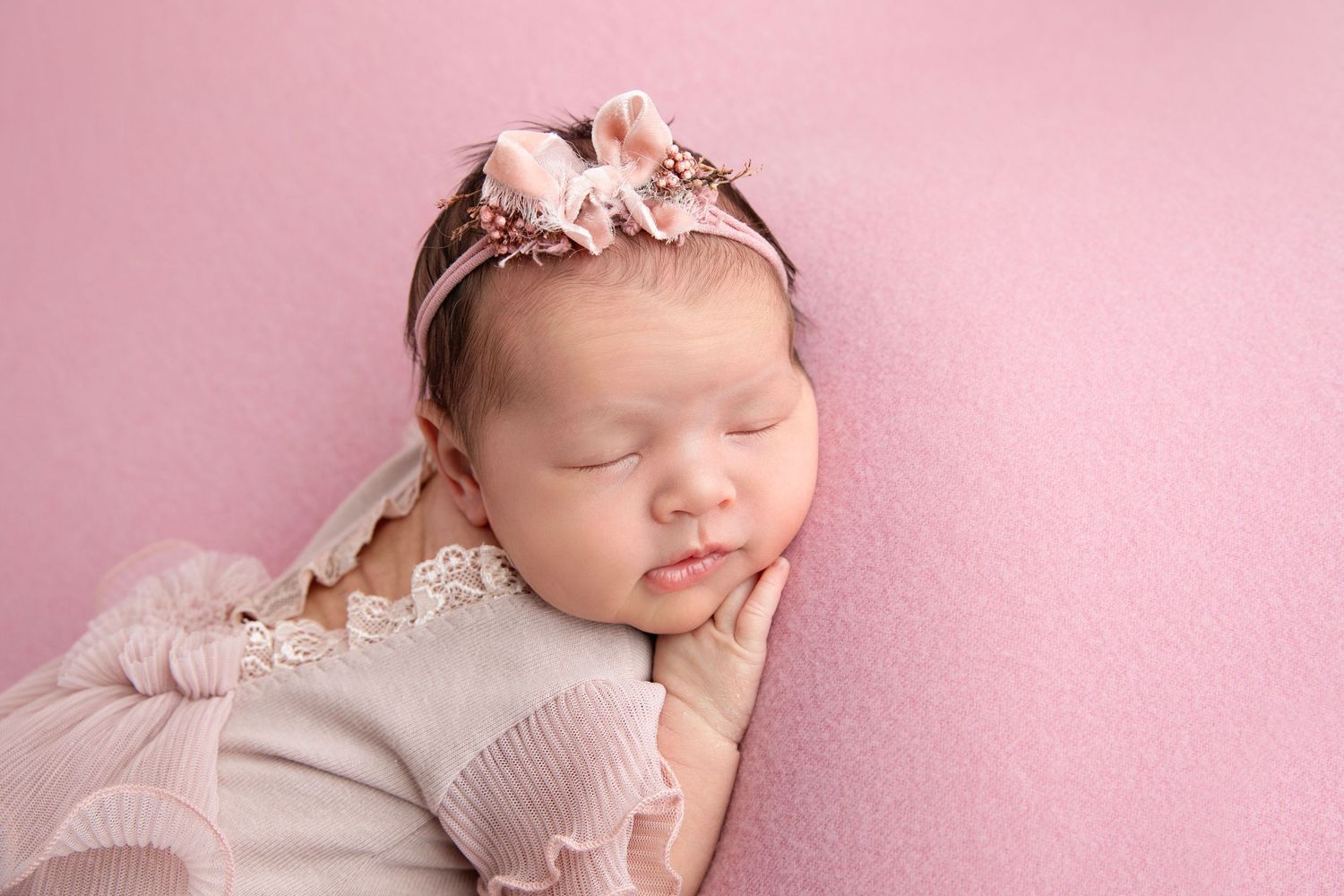 Sleeping newborn baby girl in pink outfit and floral headband on soft pink background.