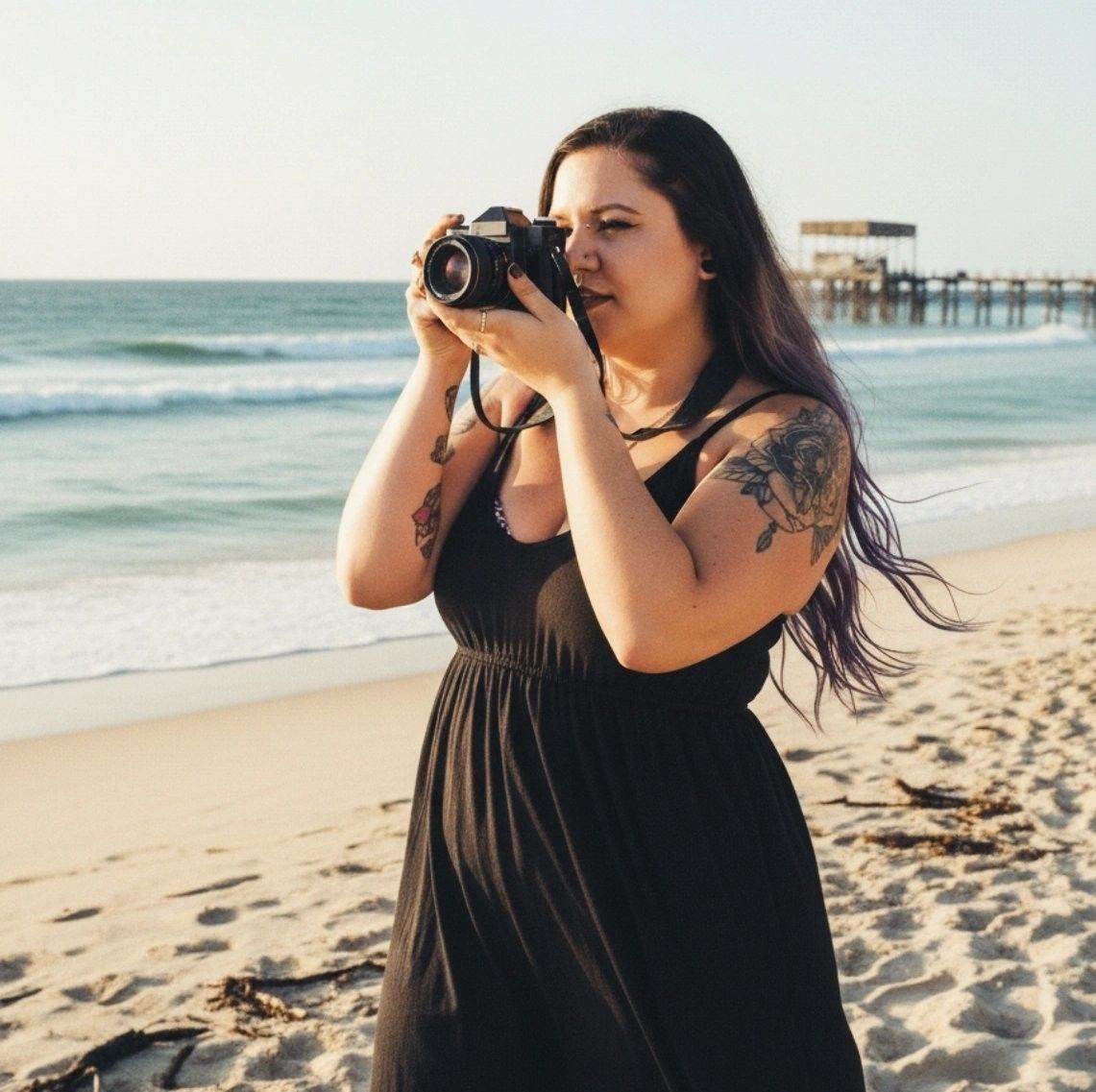 Woman with tattoos in black dress photographing on a sunny beach with a DSLR camera, pier visible in background.
