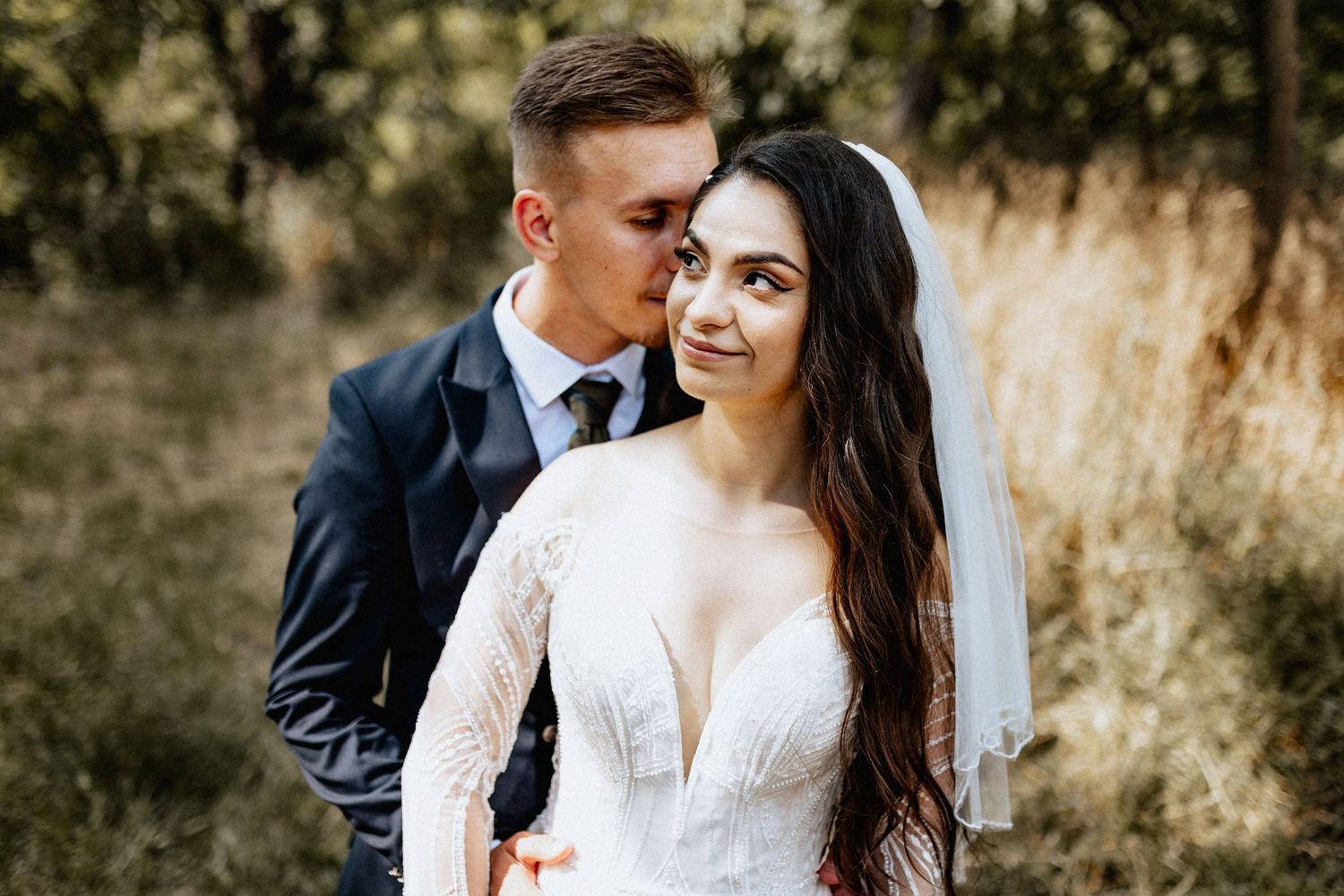 Groom in navy suit kisses bride in white lace gown and veil near a red ornate door.