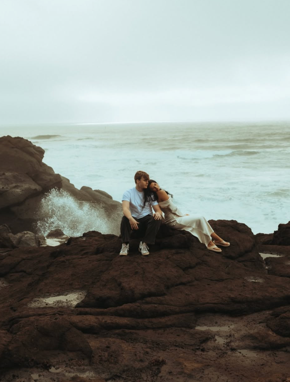 Couple sitting together on dark coastal rocks with ocean waves crashing behind them on a moody, overcast day.