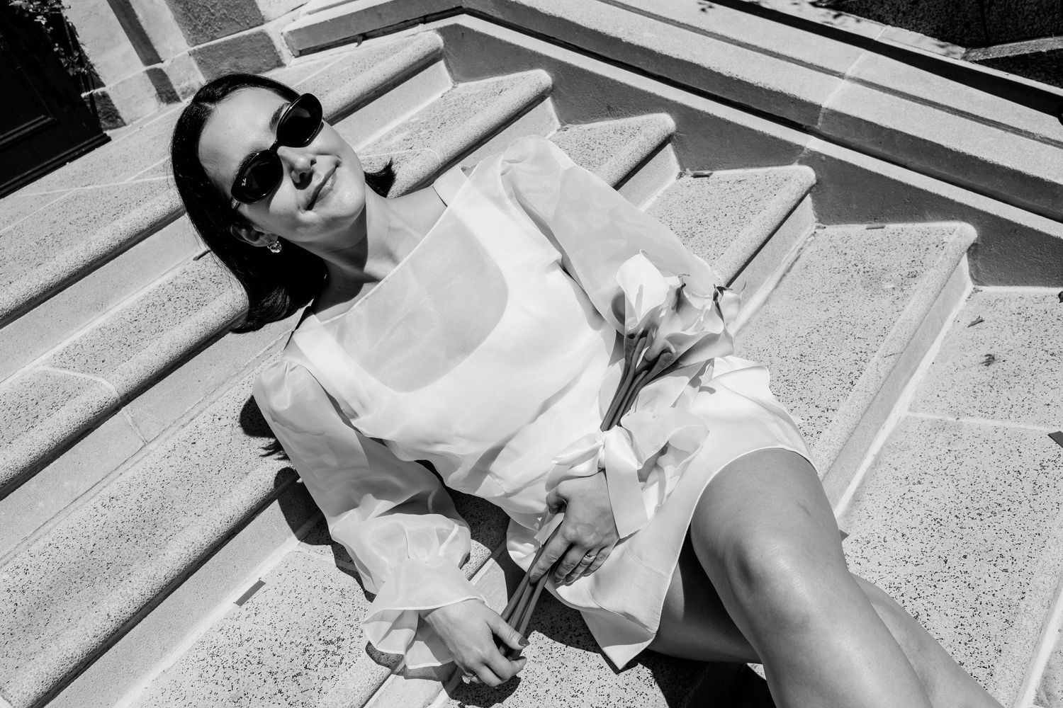 Black and white photo of a stylish woman in sunglasses and white dress relaxing on stone steps holding flowers.