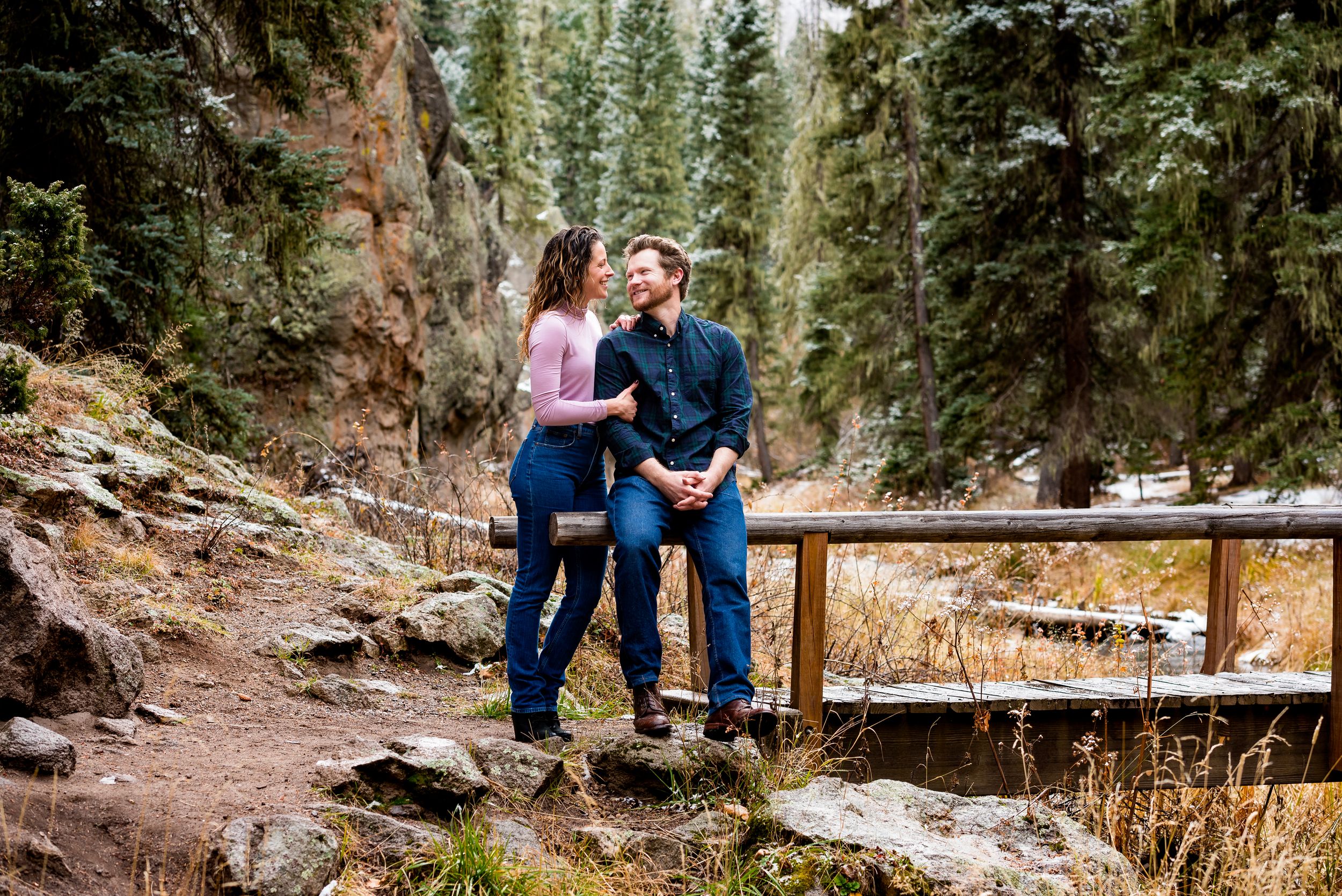 A couple shares an intimate moment on a wooden bridge surrounded by evergreen trees in a mountainous setting.