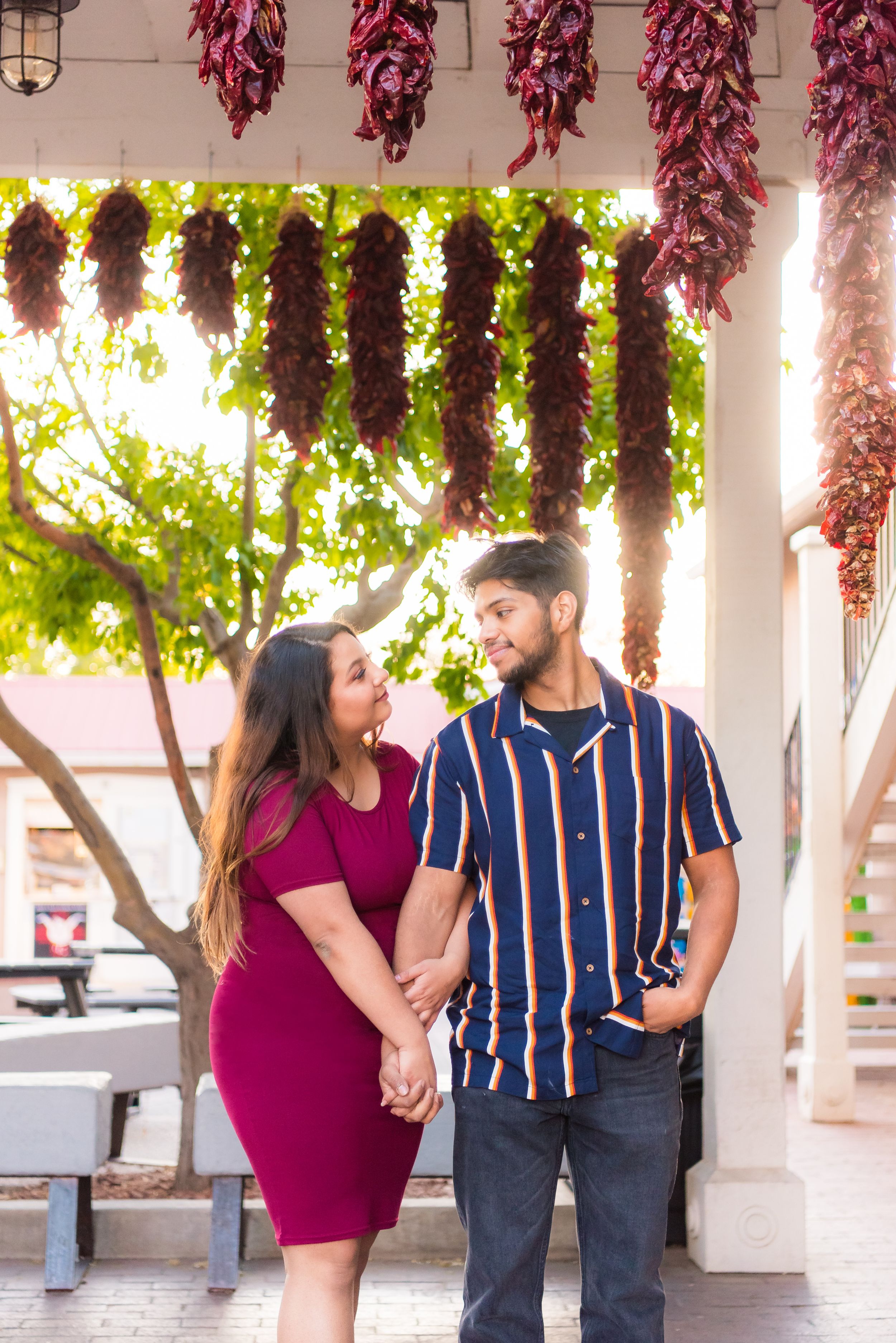 A couple poses under decorative dried red chili peppers hanging from a porch with green foliage in the background.