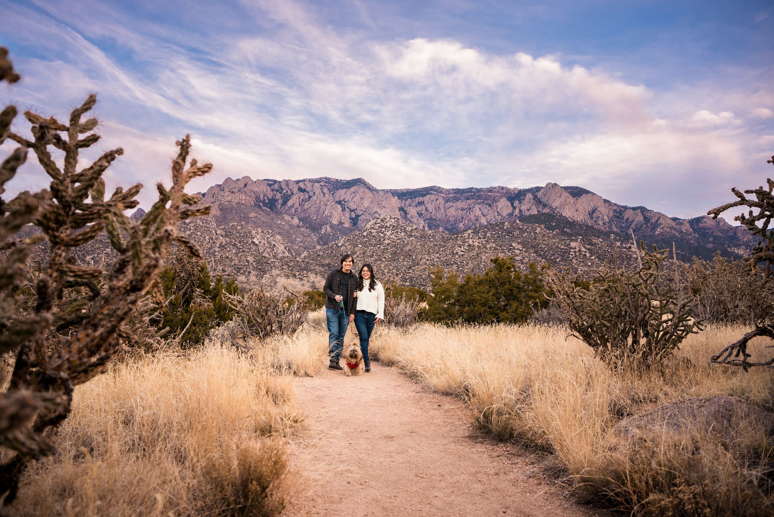 A couple standing on a desert trail with mountains and blue skies in the background.