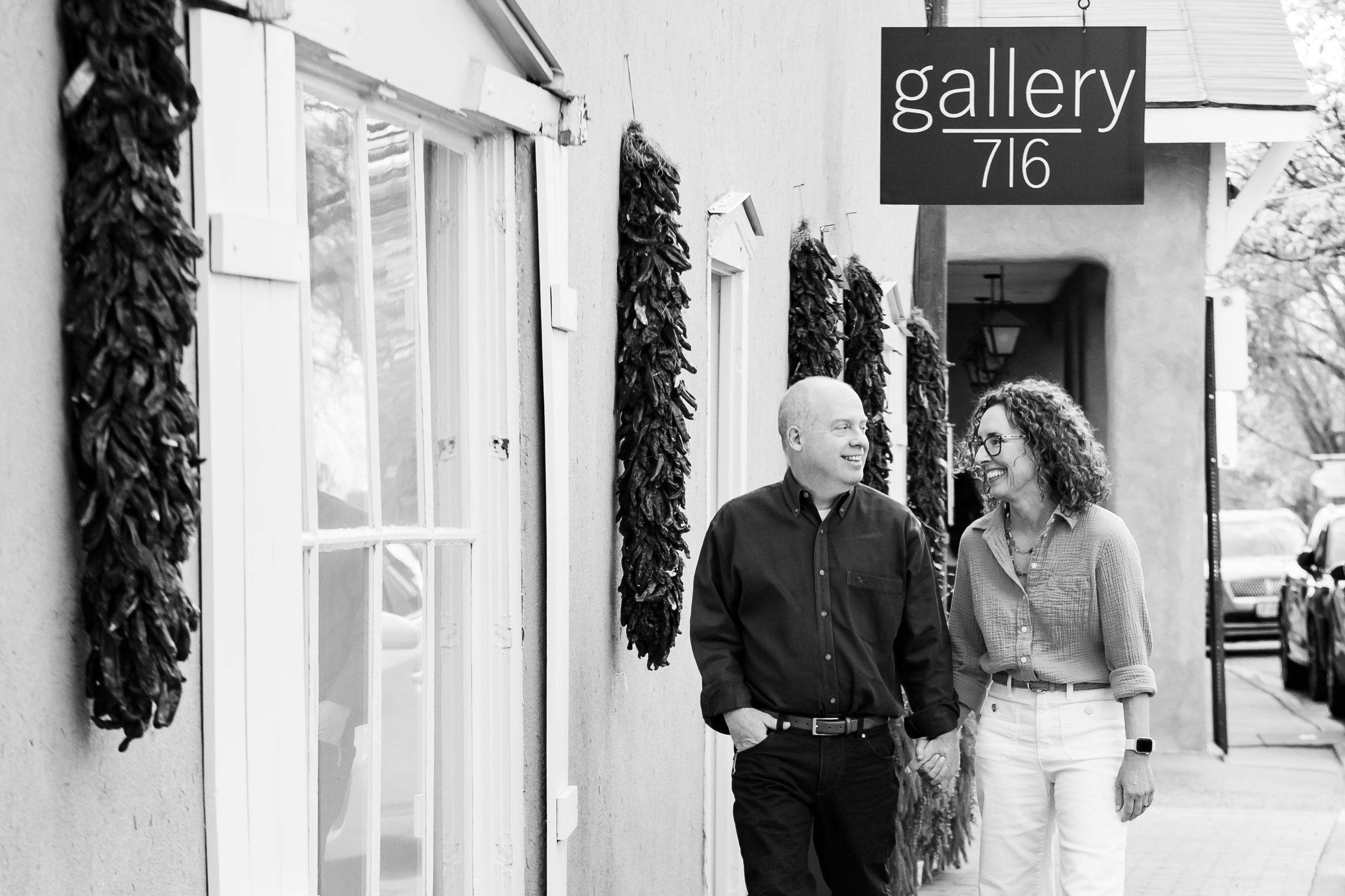 A couple walks and smiles outside Gallery 716, a Southwestern adobe building with hanging dried chili pepper ristras.