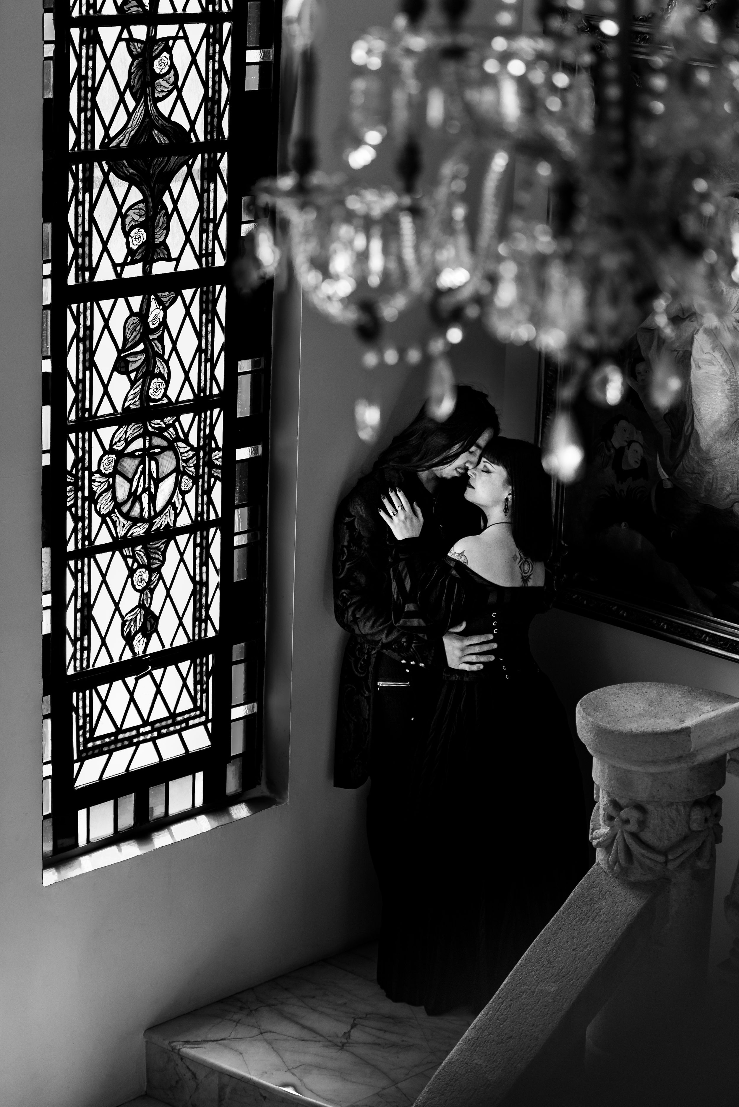 Dramatic black and white photo of a couple in dark clothing embracing near a gothic stained glass window.