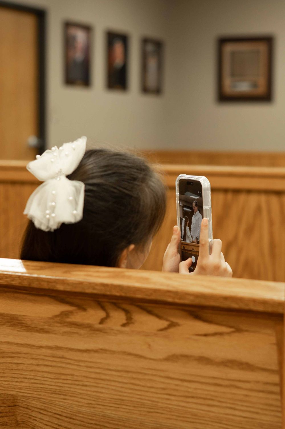 Young girl with white bow hair clip sits in wooden pew, recording ceremony on smartphone in formal meeting room.