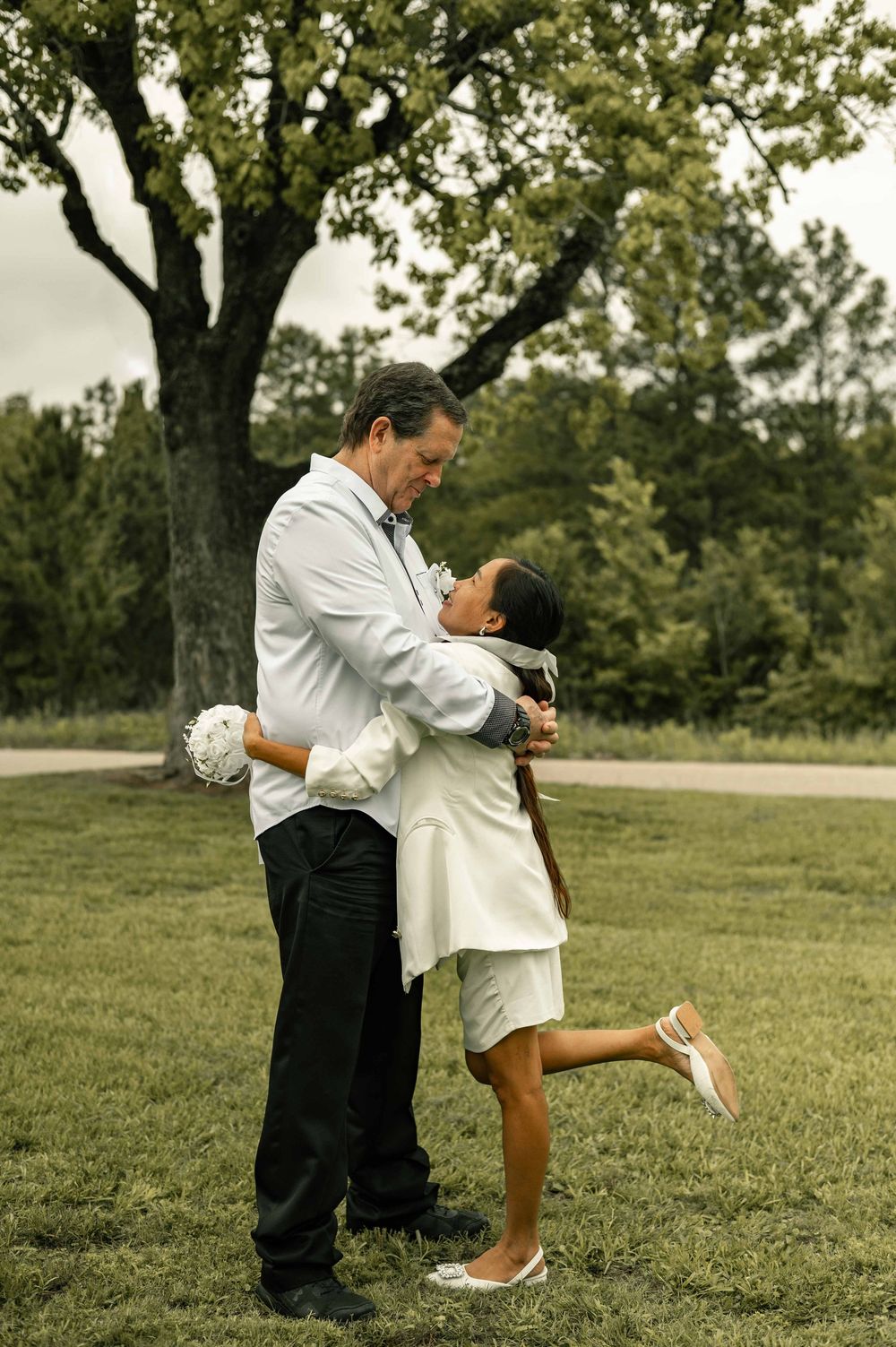 A couple shares a romantic embrace outdoors, with the bride in white lifting her leg as they lean in for a kiss near lush green trees.