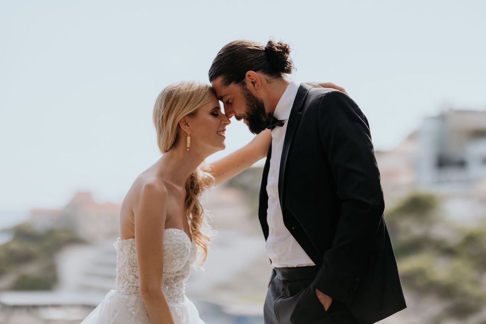 Bride and groom share a romantic forehead touch against a Mediterranean backdrop.