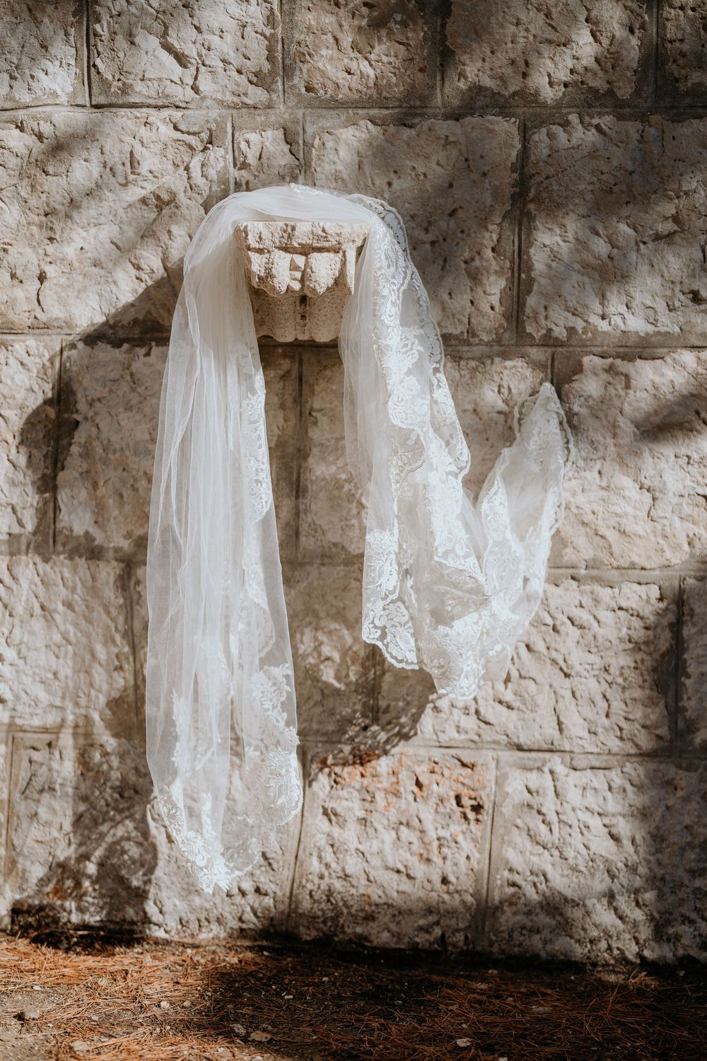 Delicate white lace bridal veil draped over ancient stone wall in sunlight.