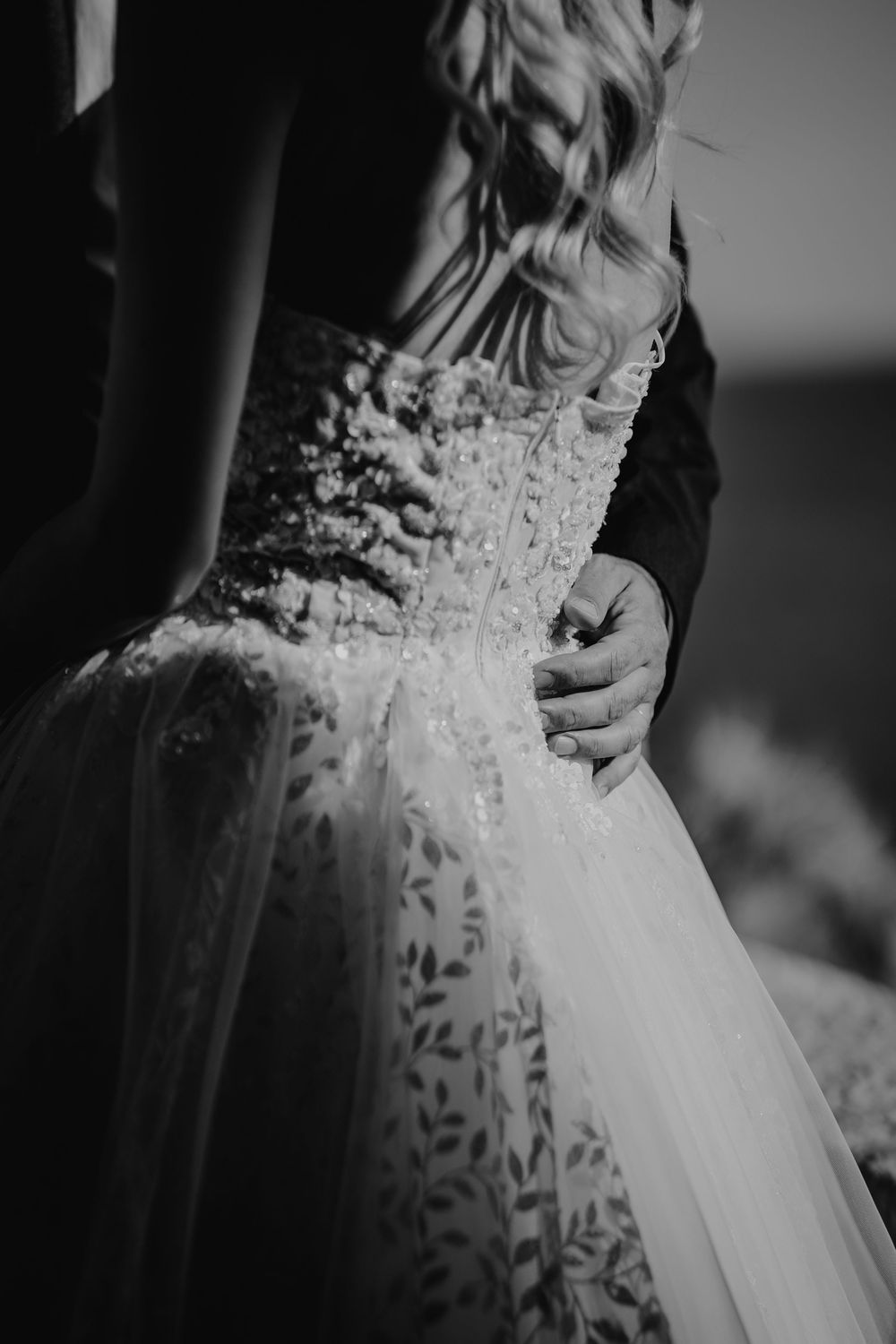 Black and white close-up of groom holding bride's embellished wedding gown waist.