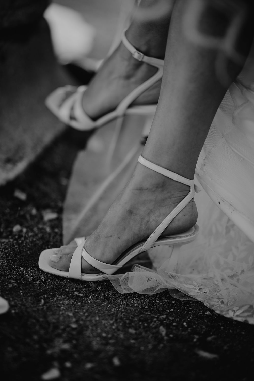 Black and white close-up of a bride wearing elegant white strappy heeled sandals with a tulle dress.