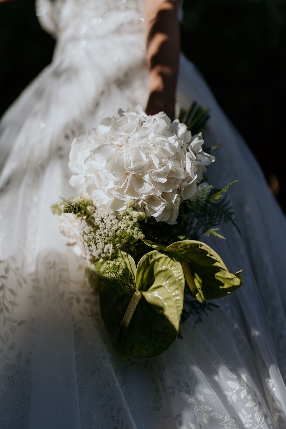 Bride holding a white hydrangea and green anthurium wedding bouquet in sunlight against lace dress.