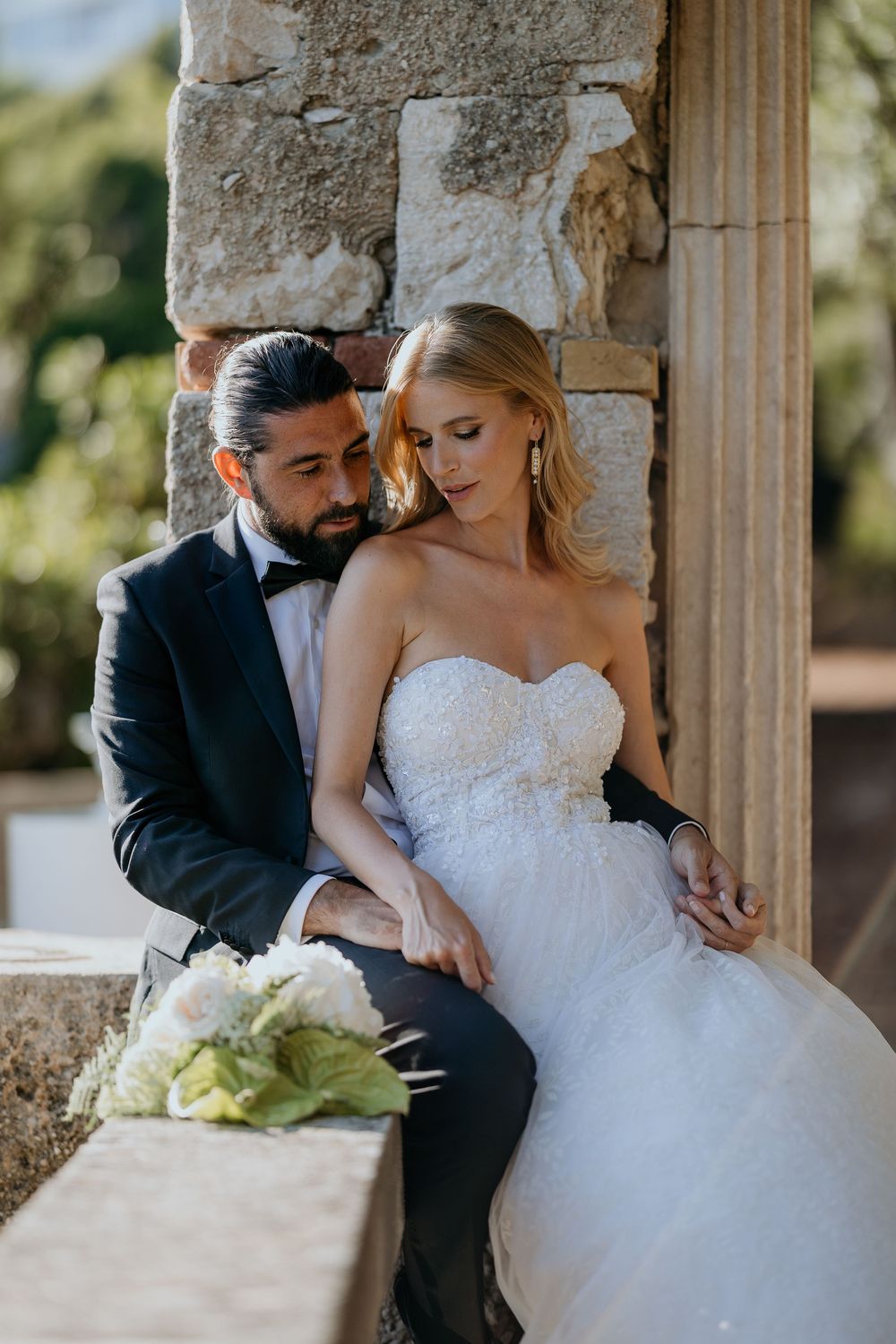 Bride and groom sitting together by stone columns, she in a strapless lace gown, he in a tuxedo.