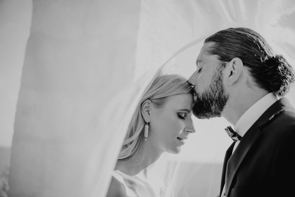 Black and white romantic photo of bride and groom nearly kissing under a flowing white veil.