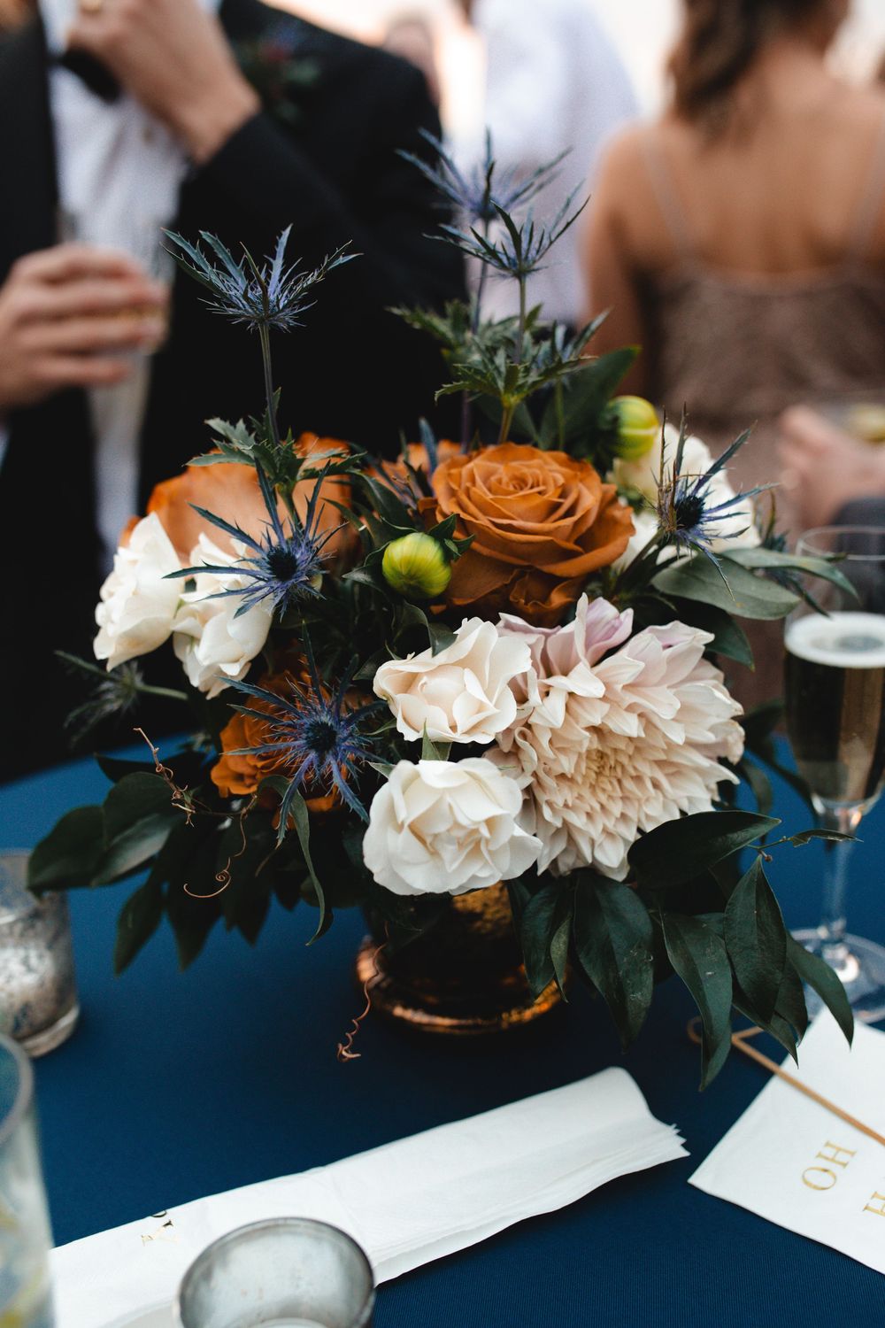 Elegant wedding centerpiece with burnt orange roses, white dahlias, and blue thistle in a copper vase on navy tablecloth.