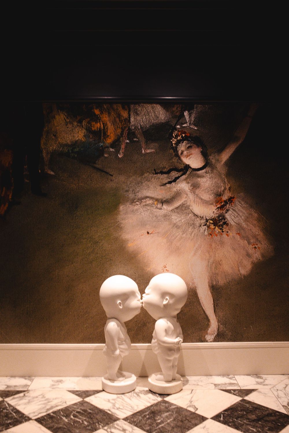 Two white kissing figurines in front of an illuminated Degas ballerina painting on a marble floor.