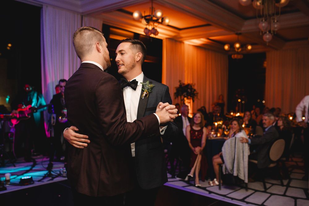 Two grooms share their first dance at an elegant wedding reception with warm lighting and live band.