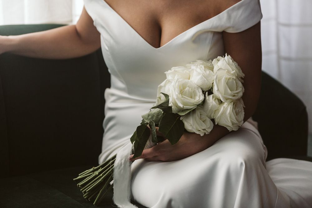 Bride in white off-shoulder gown holding elegant white rose bouquet while seated, perfect for wedding inspiration.