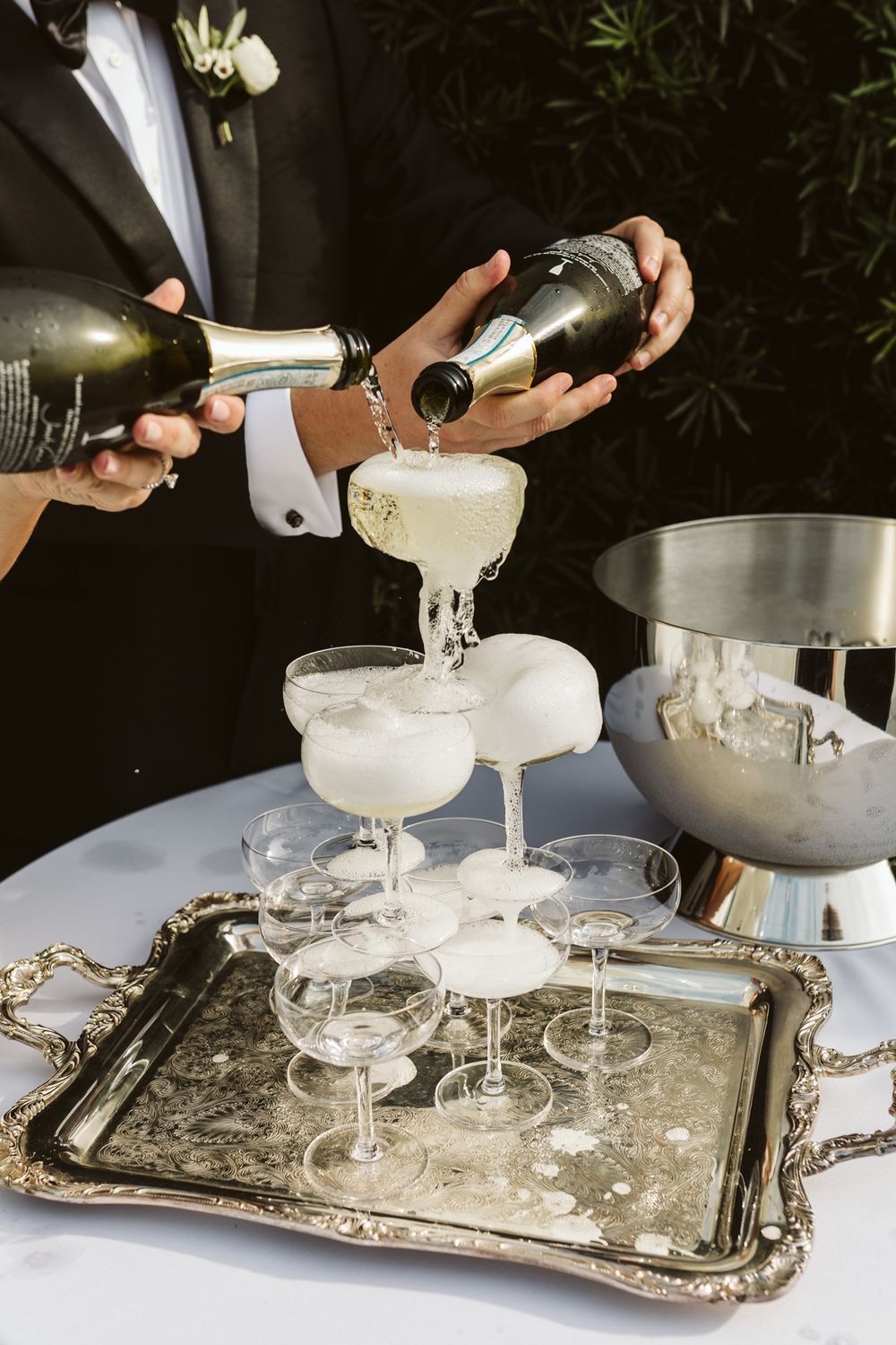 Champagne tower being filled at a wedding reception with bottles pouring bubbly into coupe glasses on silver tray.