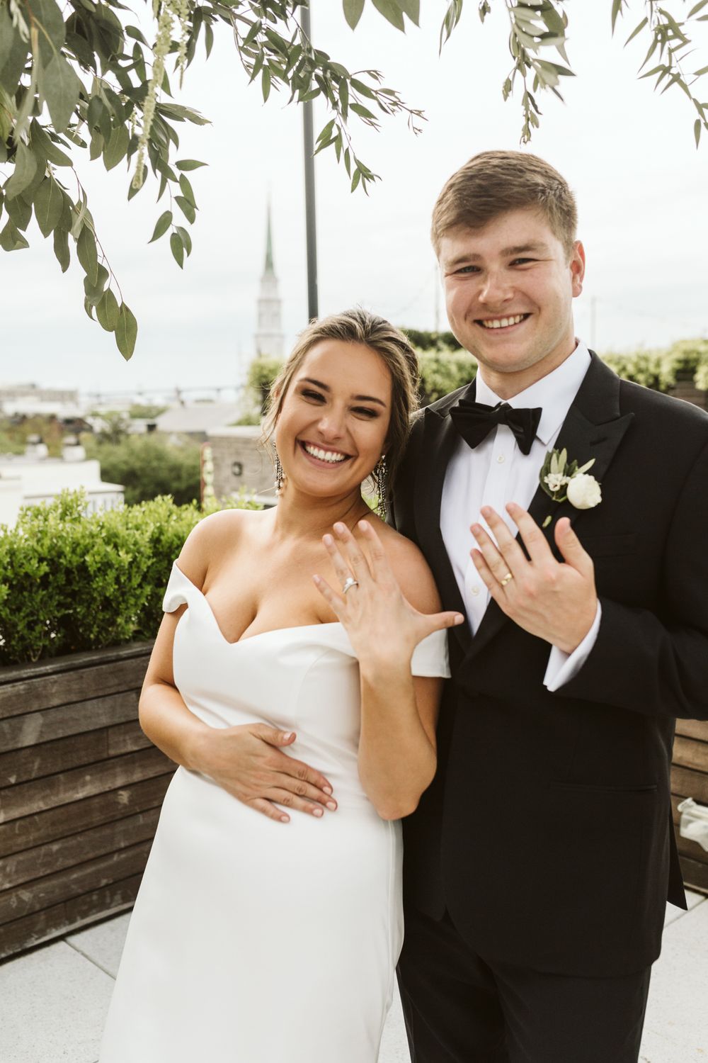 Smiling bride and groom in wedding attire showing off their rings on a rooftop venue with city views.
