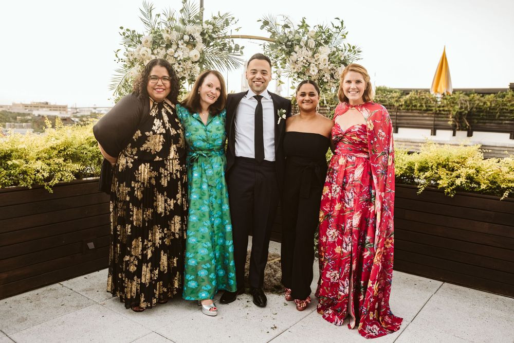 Five elegantly dressed guests smile together on a rooftop terrace at a wedding reception.