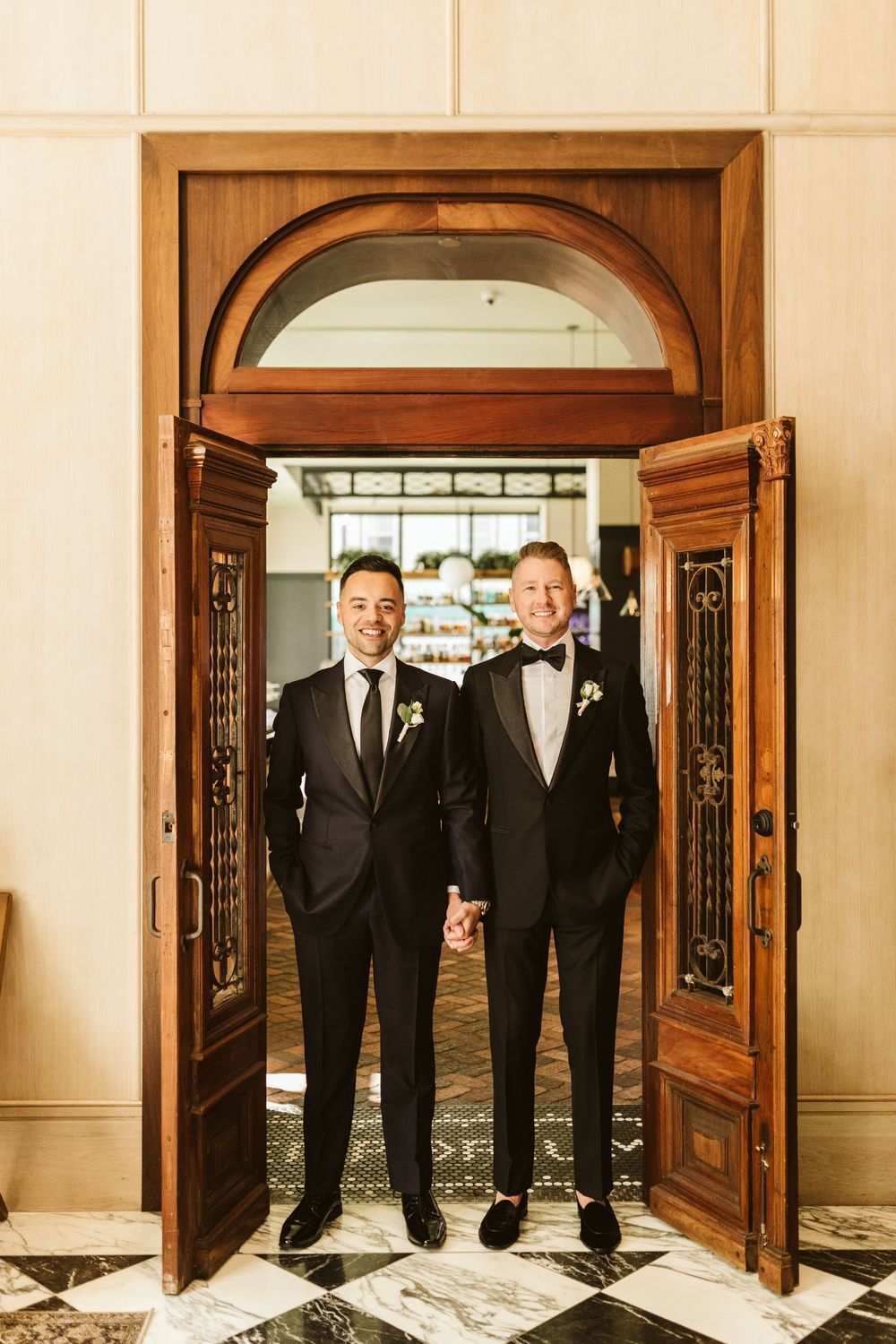 Two grooms in black tuxedos hold hands smiling in elegant wooden doorway on their wedding day.