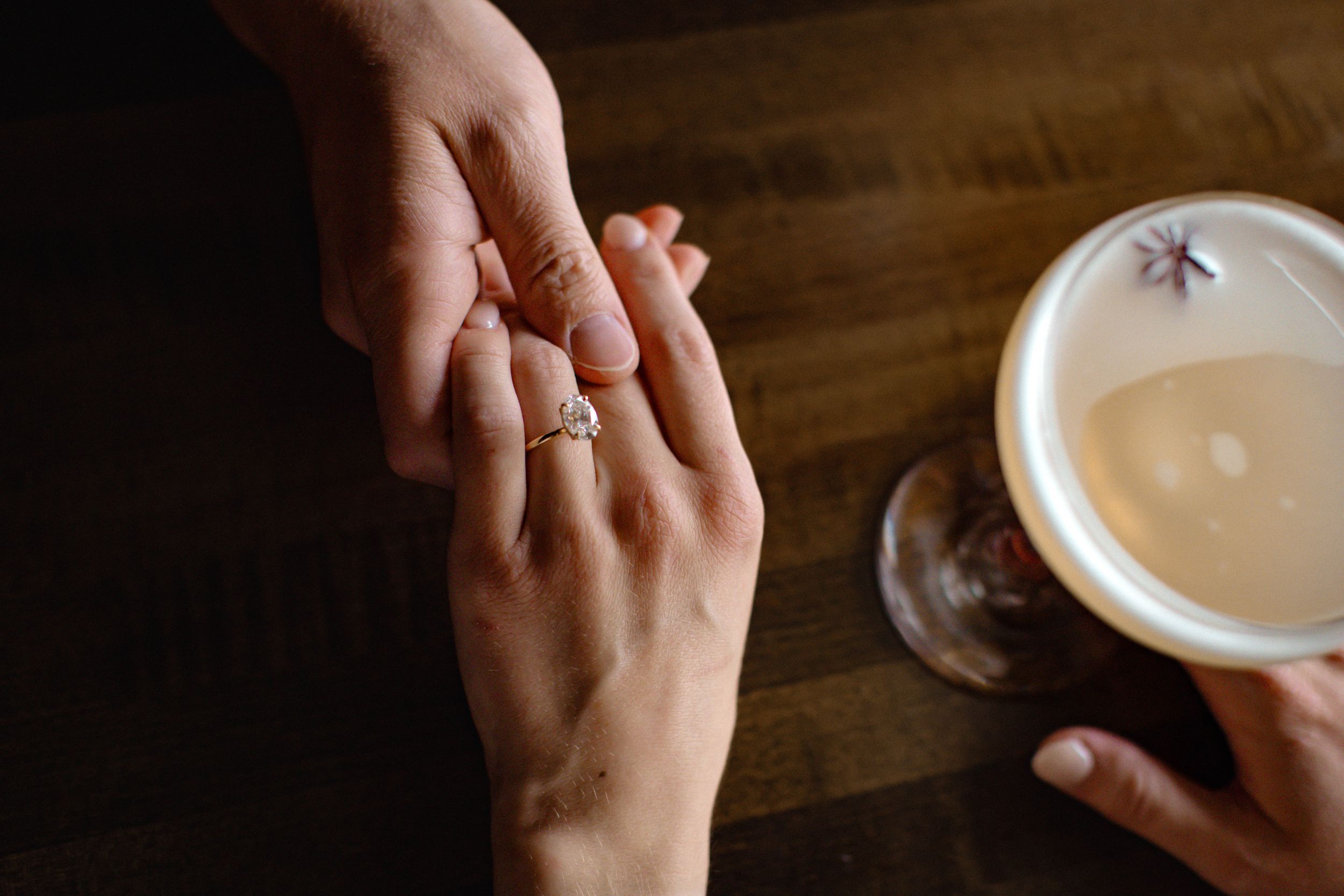 A detail shot taken from above shows a man and woman holding hands across a table with a cocktail and engagement ring visible.