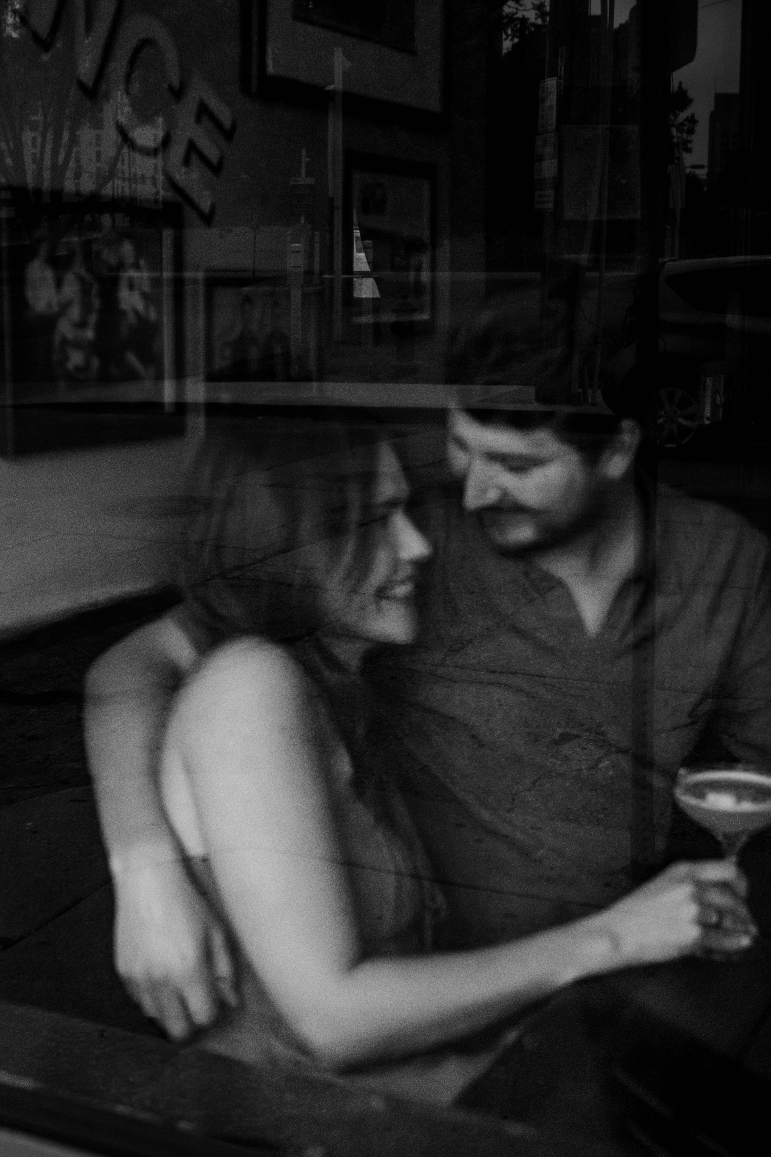 The view through a bar window as a man and woman sit close in a booth and smile at each other, his arm around her shoulders.