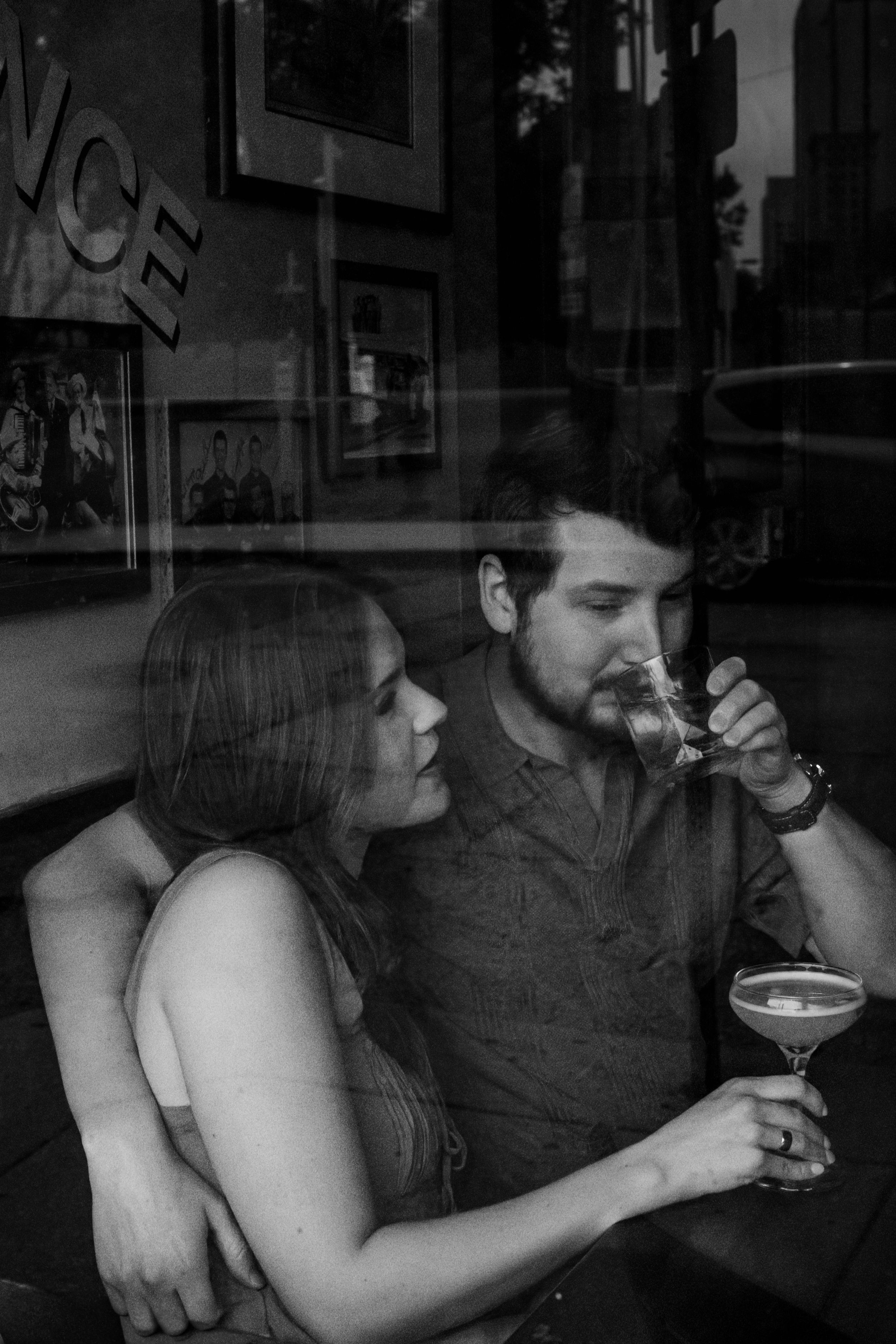 The view through a bar window as a man takes a sip of a cocktail with his arm around his fiance's shoulders as she looks up at him.