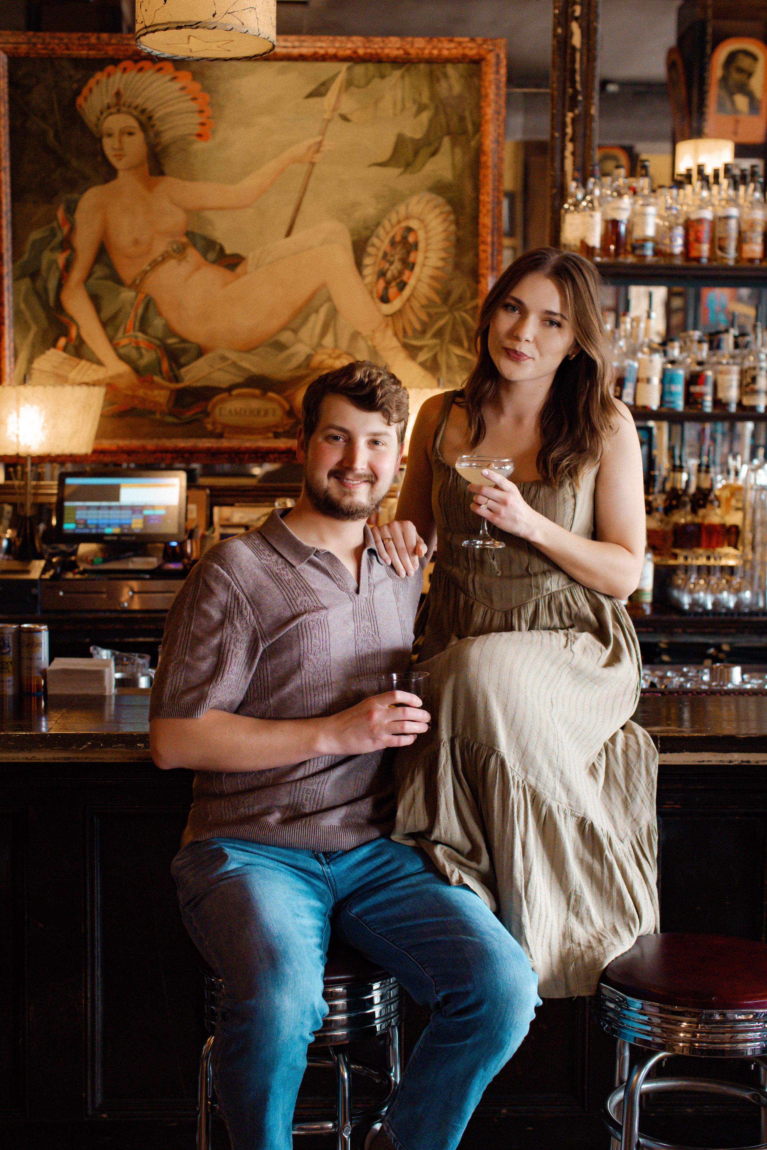 A man sits on a bar stool with a woman on the bar next to him, both holding cocktails and smiling softly at the camera.