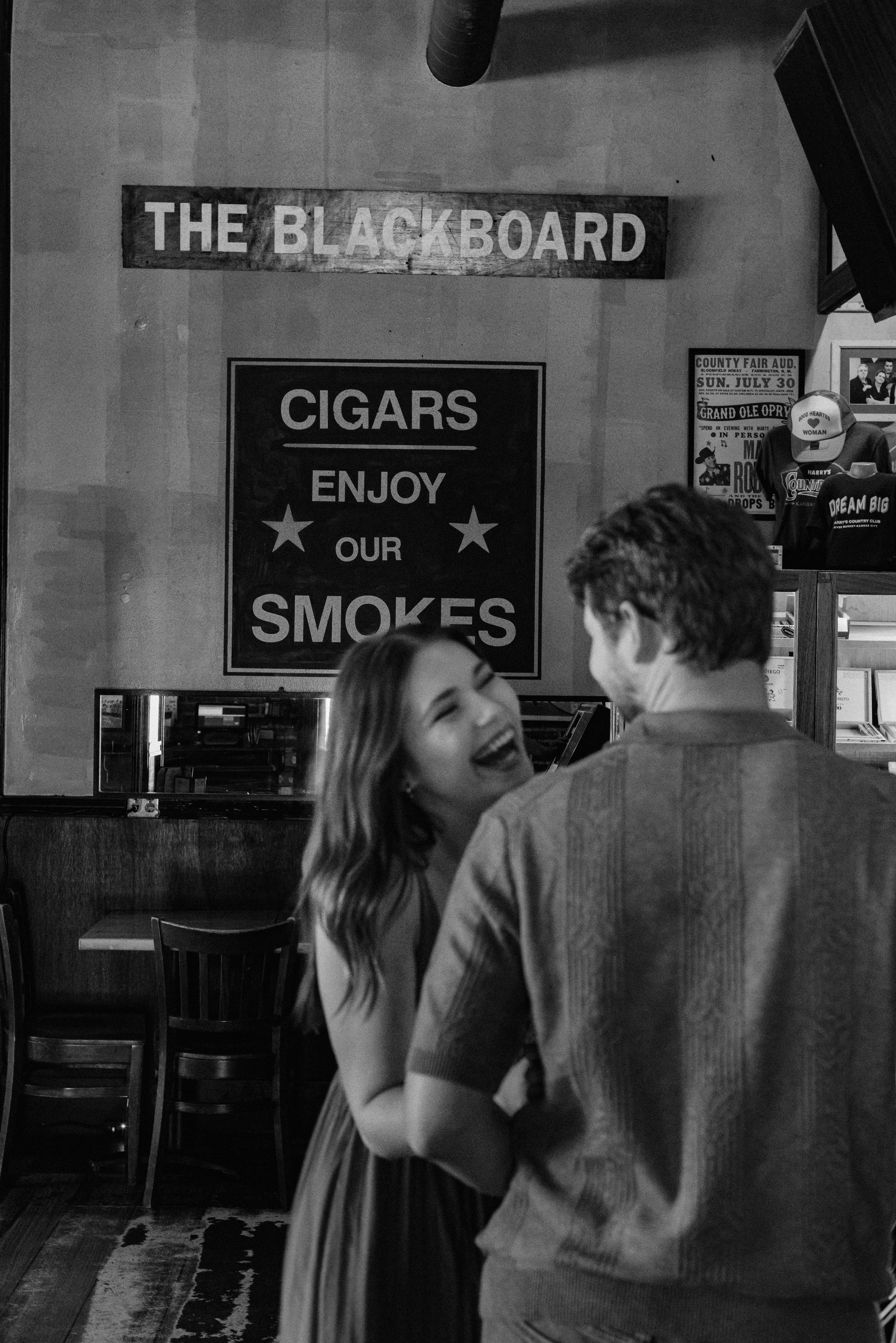 A black and white image of a woman looking at her fiance and laughing during a Kansas City engagement photo session in Harry's Country Club.