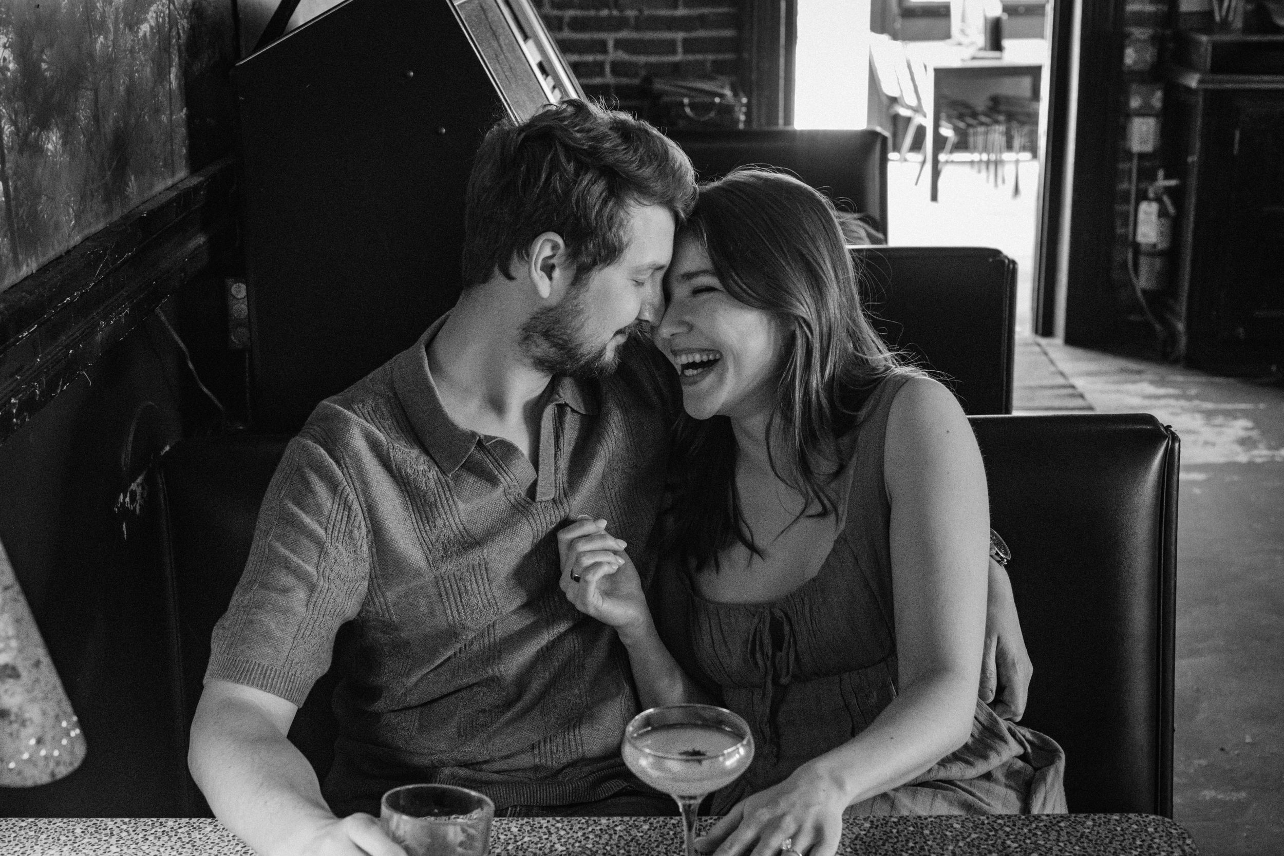 A man and woman get cozy and laugh together in a booth during a Kansas City engagement photo session at Harry's Country Club.