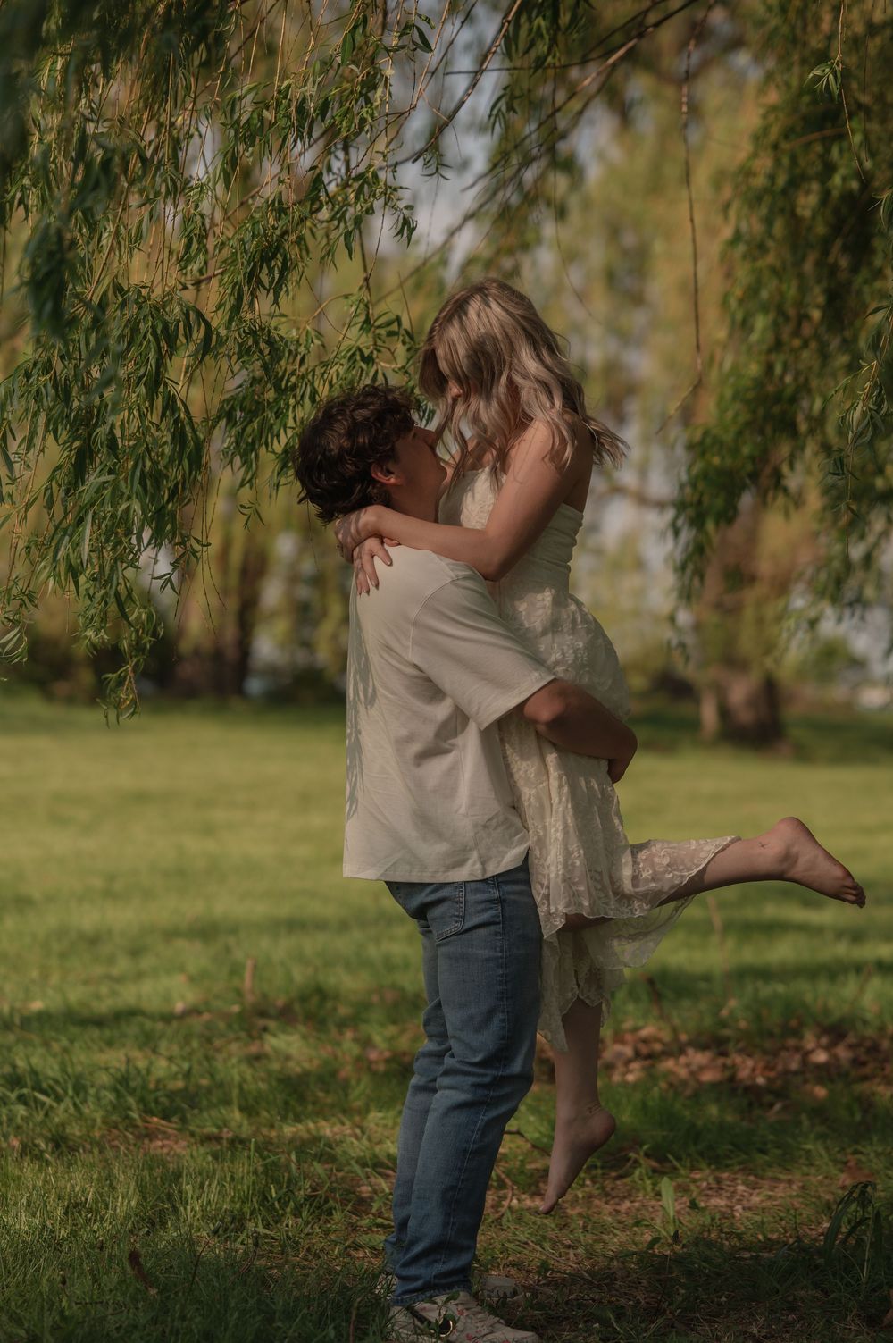 Couple sharing a romantic embrace under a weeping willow tree in a sunlit park.