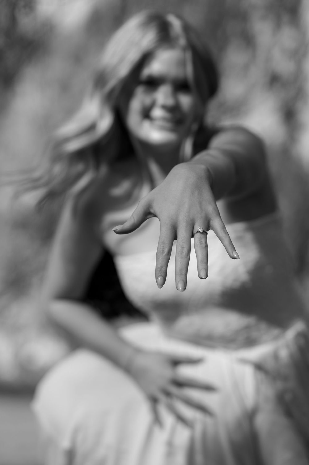 Black and white photo of smiling blonde woman showing off engagement ring on her outstretched hand toward camera.