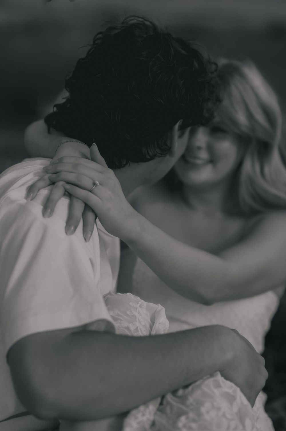 Black and white photo of a smiling bride embracing her groom, showing off her engagement ring.