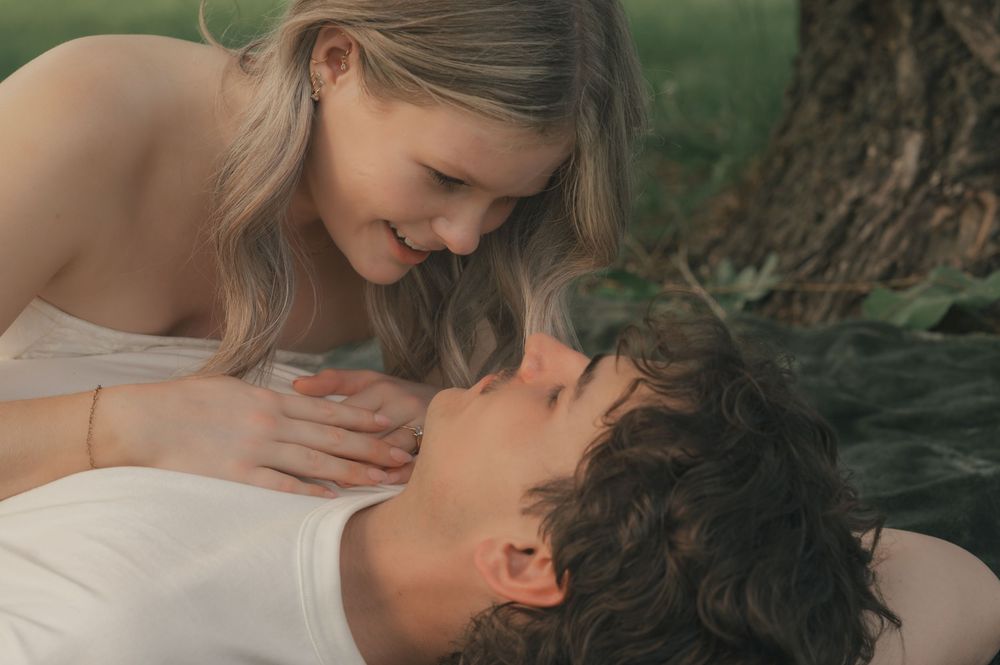 A young couple shares a tender romantic moment outdoors, with a smiling woman leaning over a man lying on the grass.