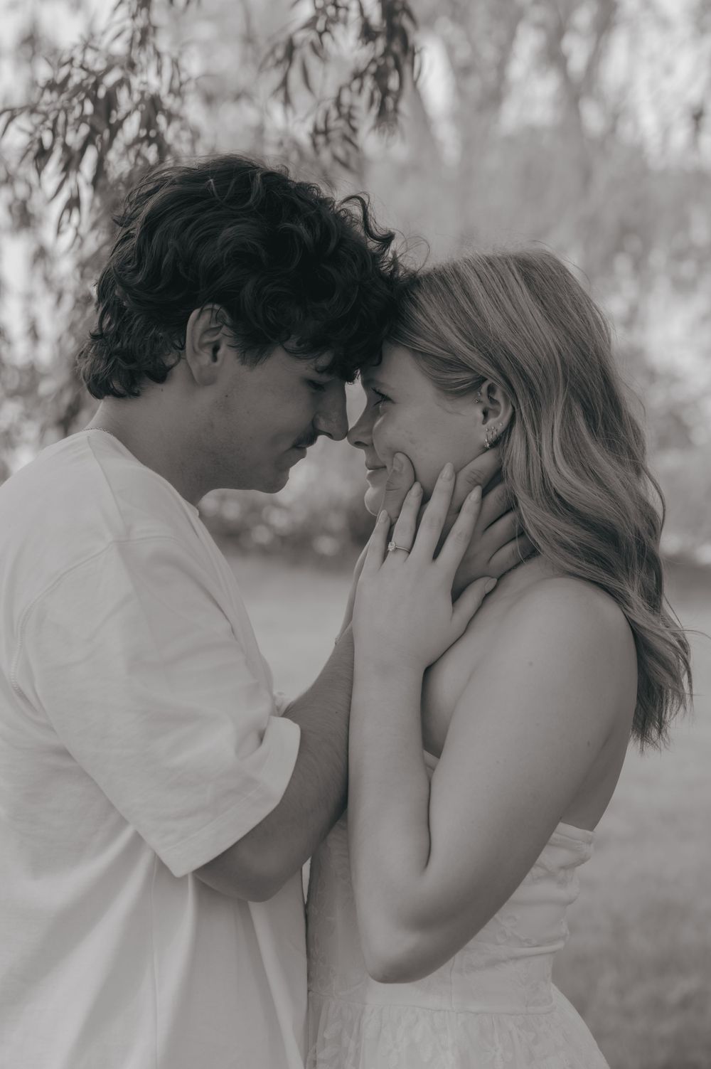 Black and white photo of a romantic couple touching foreheads and smiling outdoors under a willow tree.