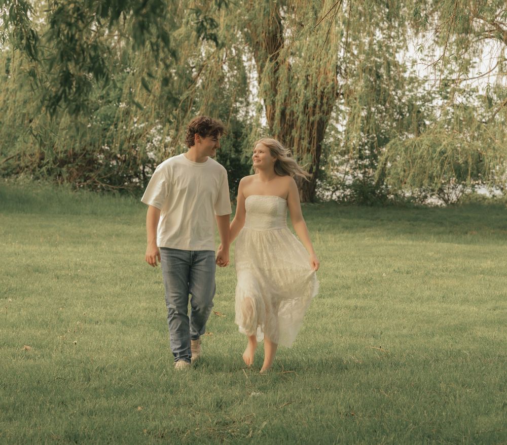 A young couple walks hand-in-hand through a lush green park, she in a white dress, he in a white tee and jeans.