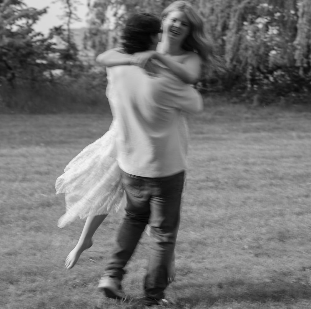 Black and white photo of a joyful couple spinning and embracing in an open grassy field.