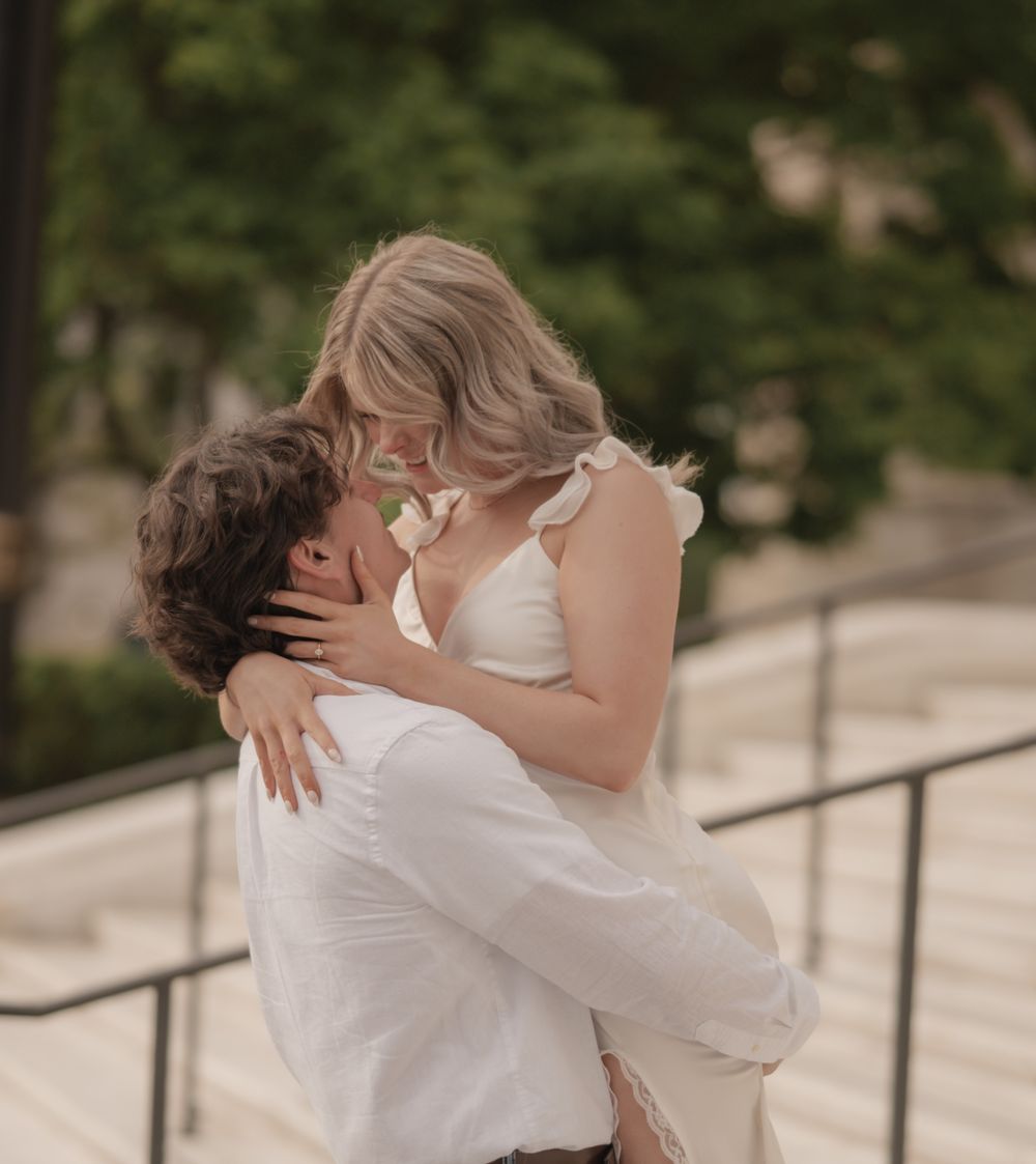 A groom lifts his bride in a romantic kiss on outdoor steps, both dressed in white wedding attire.