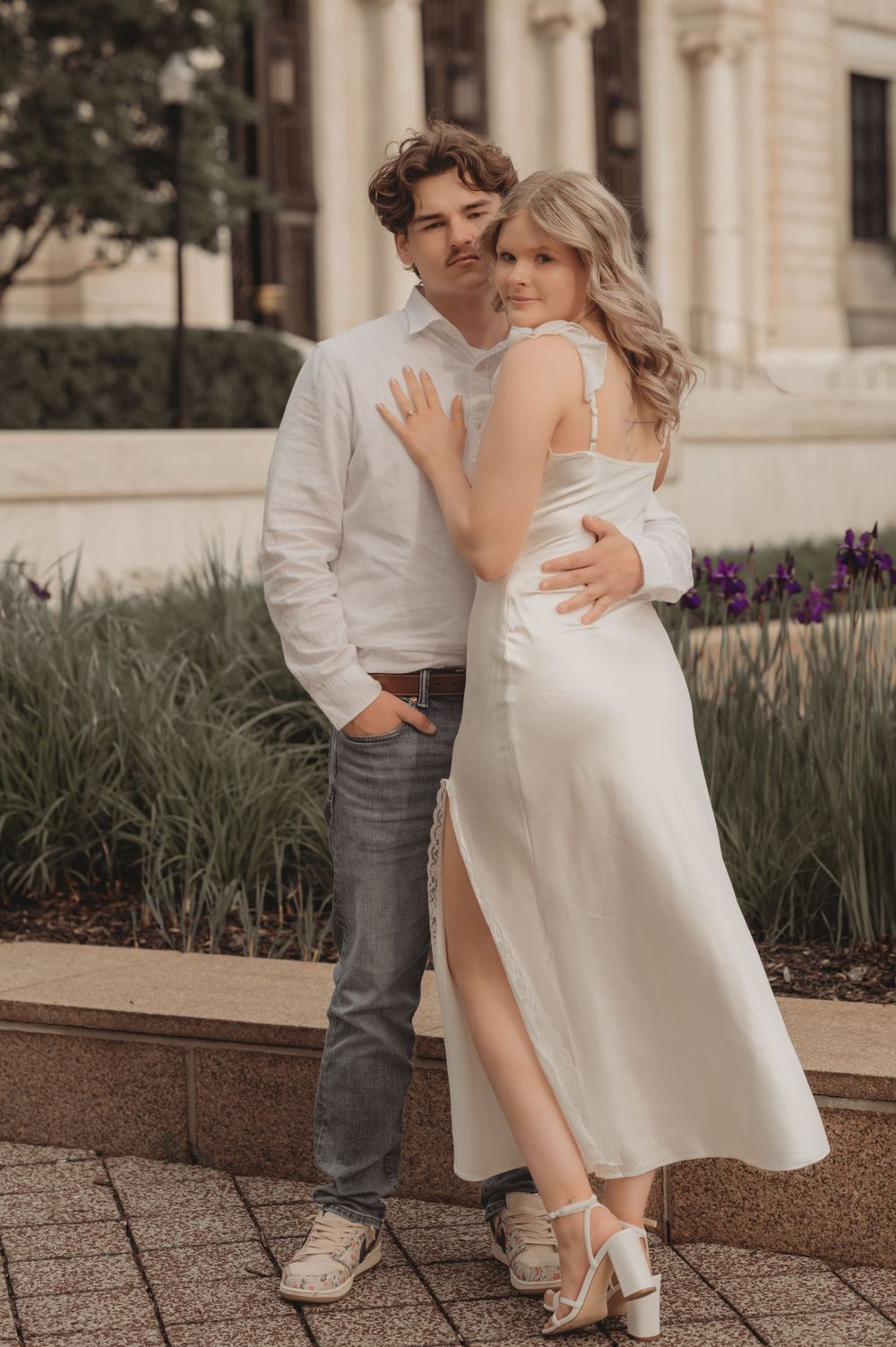 A couple in white outfits poses romantically in front of purple flowers and a stone building.