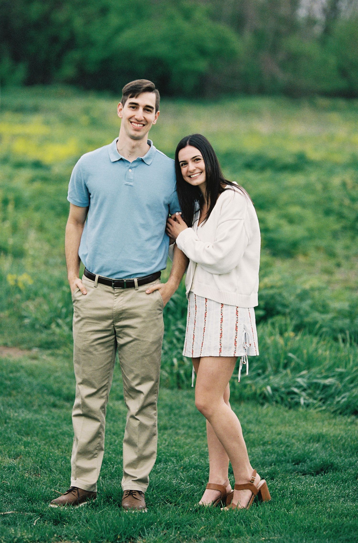 A smiling couple poses together in a green meadow, man in blue polo and khakis, woman in white jacket and striped skirt.
