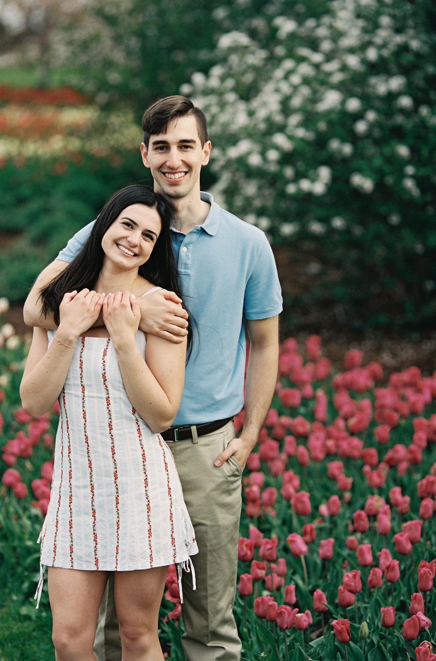 Couple embracing in front of pink tulips, woman in white dress, man in light blue polo shirt, garden setting.