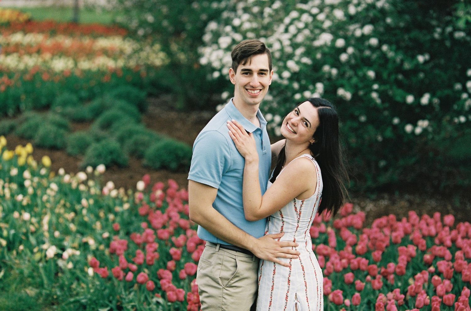 Couple embracing and smiling in a vibrant garden filled with pink and red tulips in full bloom.