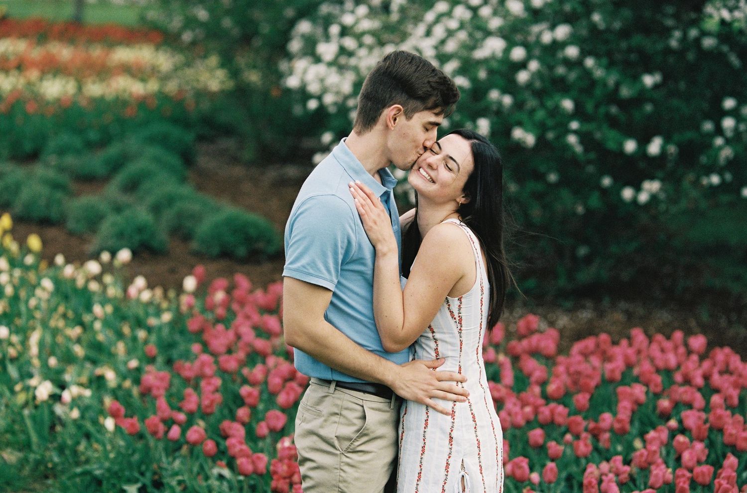 A couple embraces romantically among vibrant pink and red tulips in a lush garden, sharing a tender kiss.